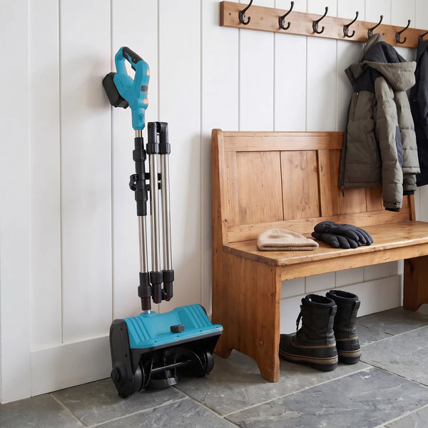 A tidy mudroom corner with an orange-and-black cordless snow shovel with its shaft folded standing upright against a white wall, a wooden bench at the right with plain winter boots, a beanie and gloves, and coat hooks above